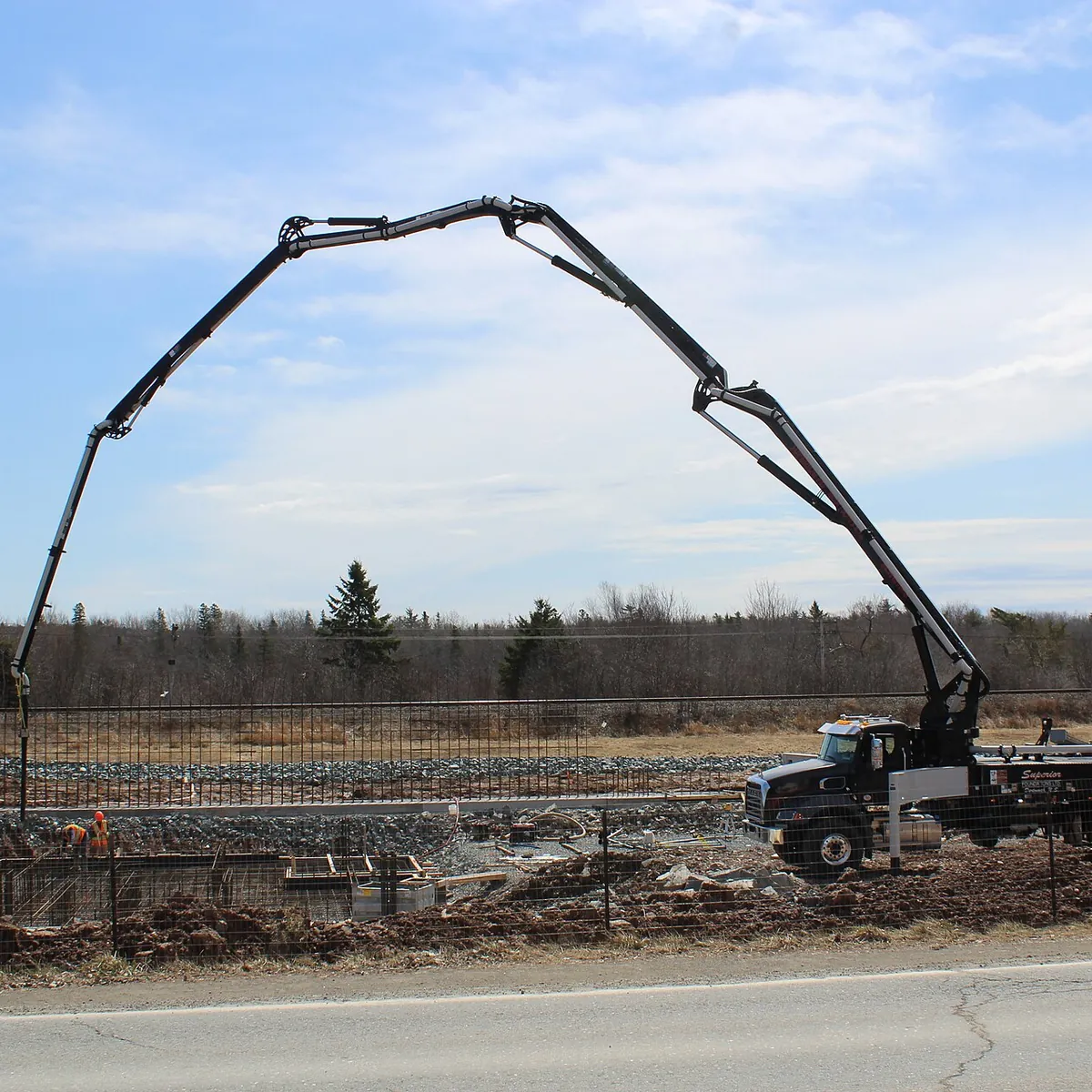 A boom pump arched over a house foundation pour, with its outriggers down beside the road